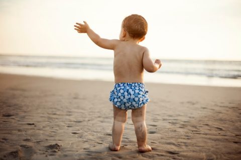 Baby standing on a beach with sandy legs in a reusable swim diaper