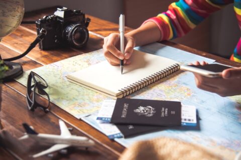 a woman in a colorful top taking notes holding a phone while planning a trip - the best travel apps